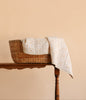 The Organic Cotton Quilt - Daffodil, featuring a cream-colored floral pattern and hand-dyed from organic cotton, is draped over the side of a woven basket on a wooden table against a plain beige background.