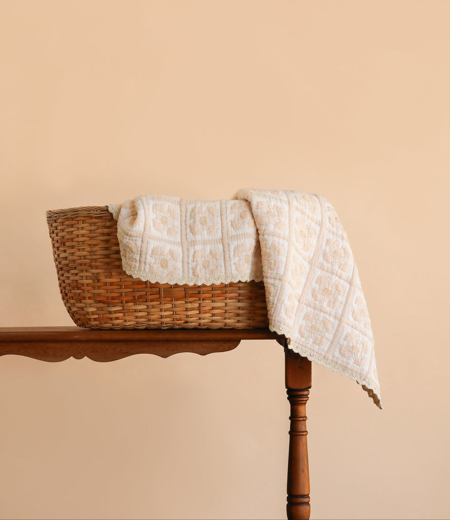 The Organic Cotton Quilt - Daffodil, featuring a cream-colored floral pattern and hand-dyed from organic cotton, is draped over the side of a woven basket on a wooden table against a plain beige background.