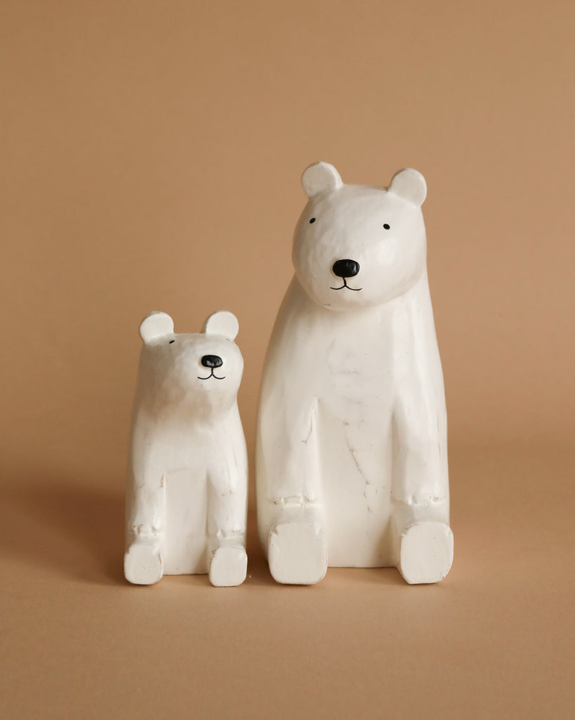 A photograph of two Handmade Tiny Wooden Polar Bears against a beige background. The larger, hand-painted polar bear sits to the right, and the smaller one sits to the left. Both bears have simple facial features and are positioned facing forward.