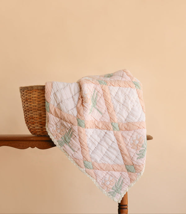 The Organic Cotton Quilt - Granny, featuring pastel patchwork and floral patterns, is draped over a wicker basket placed on a wooden table against a plain beige background.