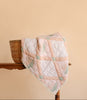 The Organic Cotton Quilt - Granny, featuring pastel patchwork and floral patterns, is draped over a wicker basket placed on a wooden table against a plain beige background.