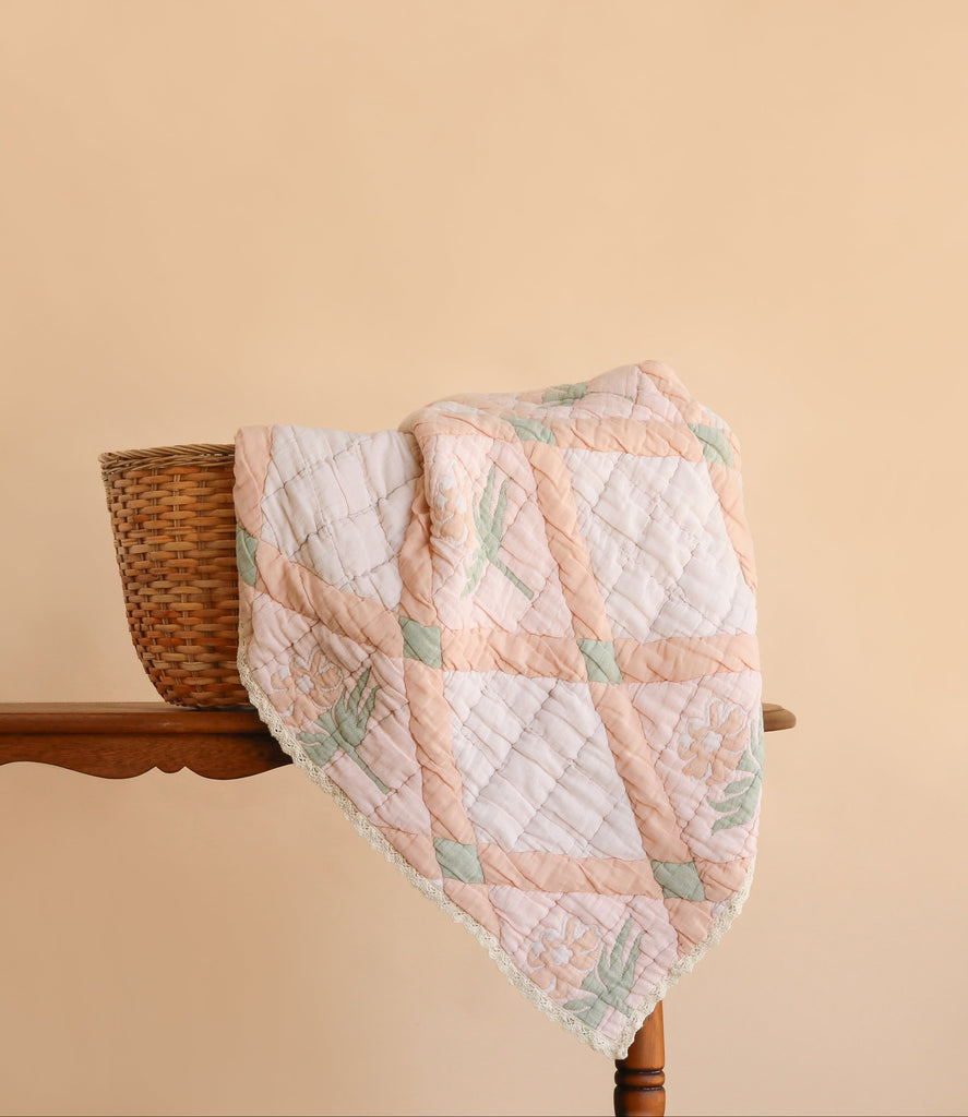The Organic Cotton Quilt - Granny, featuring pastel patchwork and floral patterns, is draped over a wicker basket placed on a wooden table against a plain beige background.