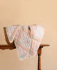 The Organic Cotton Quilt - Granny, featuring pastel patchwork and a delicate lace edge, is draped over a wicker basket on a wooden table against a light beige background.