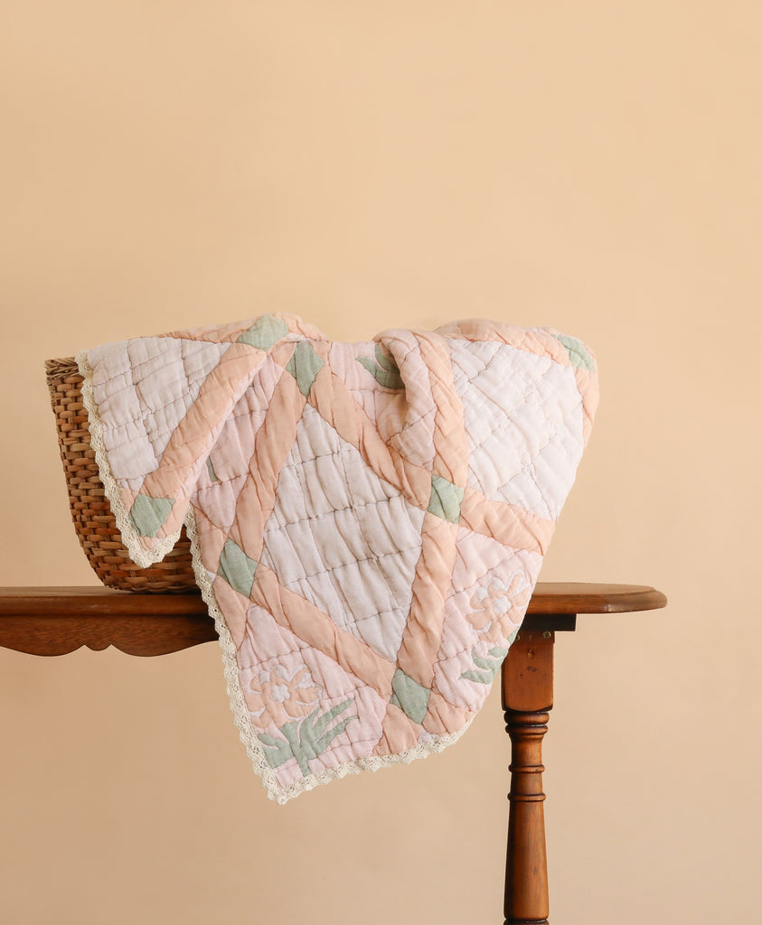 The Organic Cotton Quilt - Granny, featuring pastel patchwork and a delicate lace edge, is draped over a wicker basket on a wooden table against a light beige background.