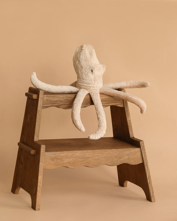 The Senger Naturwelt Cuddly Animal - Octopus, made from organic cotton with long floppy arms, rests on a wooden step stool against a beige background—an ideal calming companion for cozy moments.