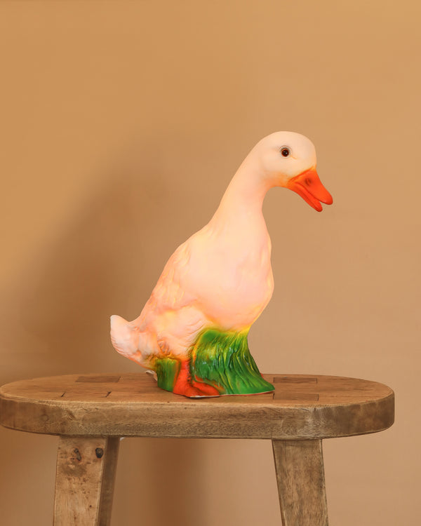 The Duck Lamp, with its orange beak and green grass base, adds a whimsical touch to any child's room—displayed here on a rustic wooden stool against a plain beige background.