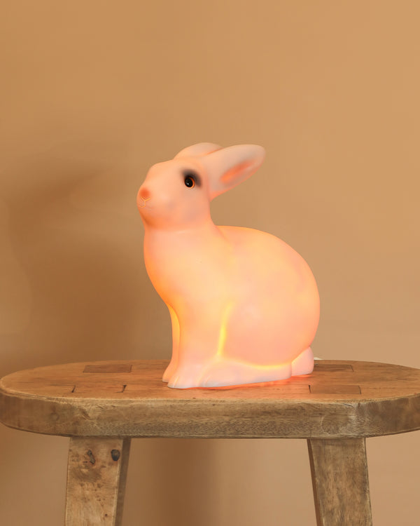 The White bunny Lamp, with its smooth white surface and adorable features, sits on a rustic wooden stool against a beige background, casting a soft, warm glow as a whimsical night light for any nursery.