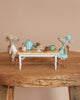 Two Maileg Mice from the Tea Party Set sit at a small wooden table adorned with a teal tea set, cups, and pastries. They wear flowers on their heads and enjoy a tea party against a beige background.