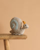 The Steiff Snaily Snail Stuffed Plush Animal, 8 inches, with a light gray body, yellow antennae, and blue spiral shell, sits on a wooden stool against a plain beige background.