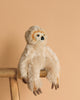 The Steiff Miguel Baby Dangling Sloth Plush Toy, 13 inches, with premium beige fur, sits on the edge of a wooden stool against a plain light peach background.