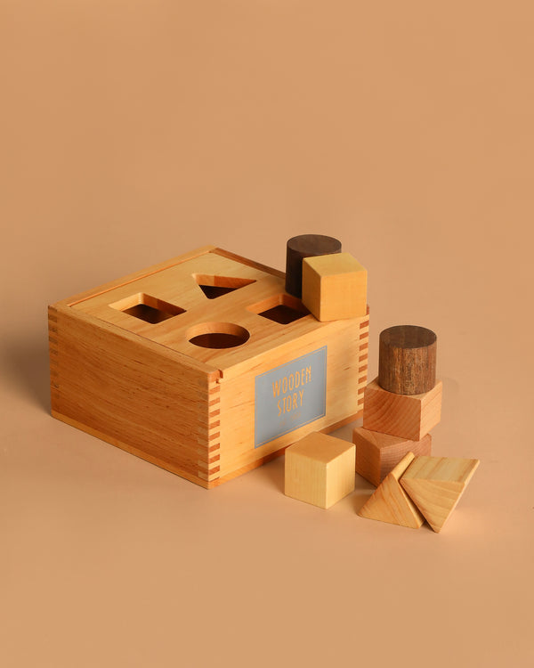 The Natural Shape Sorter Box, a wooden toy, showcases various geometric shapes like cubes, cylinders, and triangles arranged around and atop the eco-friendly box against a beige background.