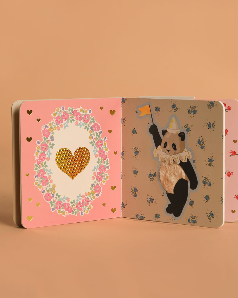 The Konges Sloejd Touch and Feel Book, made with FSC-certified materials, is open to a pink page with a gold heart in a floral wreath and another page showing a panda in a hat holding a flag amid heart and flower patterns.