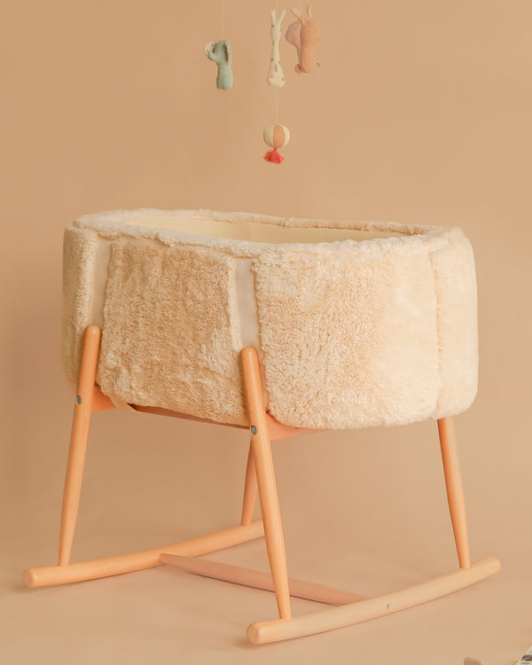 A soft, beige Charlie Crane KUKO 'Fur' Moses Basket with wooden rocker legs rests on a matching background. Plush animal mobiles hang above, creating a cozy, neutral-toned nursery scene.