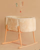 A soft, beige Charlie Crane KUKO 'Fur' Moses Basket with wooden rocker legs rests on a matching background. Plush animal mobiles hang above, creating a cozy, neutral-toned nursery scene.