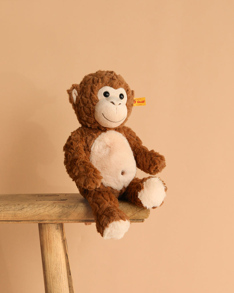 The Bodo Monkey Plush Stuffed Toy, 12 Inches, in brown and white with a stitched smile, sits on a wooden bench against a beige background. A yellow tag in its ear marks it as an authentic Steiff product.