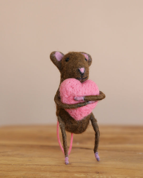 The Felt Mouse With Heart, hand-felted in India, features brown fur and pink accents on its ears, hands, and feet. It holds a pink heart while standing on wooden surface against a neutral beige background—ideal for gentle play or display.