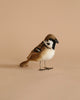 A Sparrow Bird Stuffed Animal with realistic features and brown, white, and black plumage stands on a plain beige background, displaying its profile with a sharp, black eye mask.