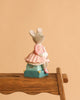 The Moulin Roty Money Box - The Little School of Dance features a small hand-drawn mouse in a pink dress holding a white bird, sitting on a teal box atop a wooden chair—perfect for decorating children's bedrooms.