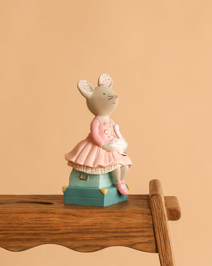 The Moulin Roty Money Box - The Little School of Dance features a small hand-drawn mouse in a pink dress holding a white bird, sitting on a teal box atop a wooden chair—perfect for decorating children's bedrooms.