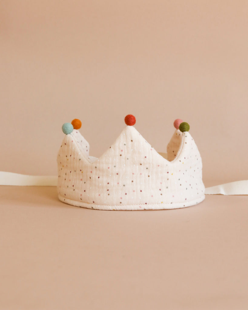 A soft, white handmade Confetti Reversible Birthday Crown adorned with small, colorful pom-poms at each of its peaks sits against a beige background. This reversible accessory features a subtle polka dot pattern and is crafted from organic cotton, with white ribbon ties extending from the sides.