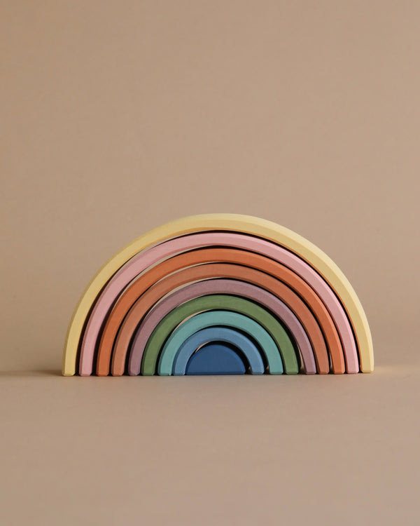 A stack of colorful wooden Raduga Grez 9-Piece Rainbow Stacker crafted from linden wood in graduated sizes arranged against a beige background. The colors, painted with non-toxic water-based paint, transition smoothly from pink to blue.