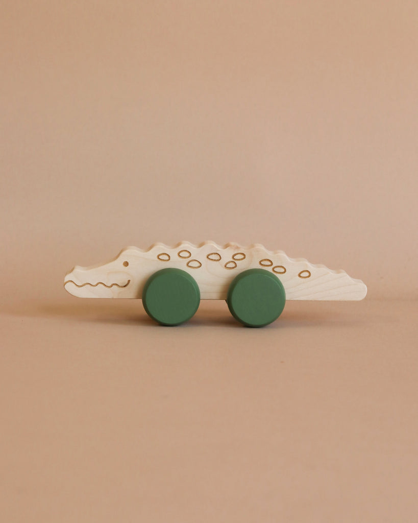 A handmade in Poland wooden toy shaped like a crocodile with simple painted details is displayed against a light brown background. This Wooden Crocodile Push Toy features two large green wheels attached to the bottom, making it perfect for helping toddlers develop fine motor skills.