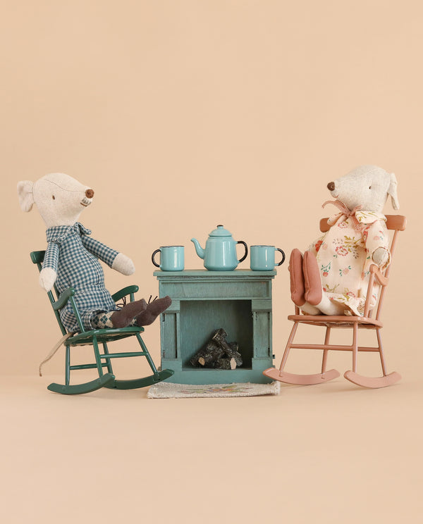 Two Maileg Grandma and Grandpa mice from the Tea Time Set relax in their rocking chairs by a miniature fireplace and table, which is set with blue teacups and a teapot against a beige background. Surrounded by adorable Maileg accessories, Grandma wears floral pajamas while Grandpa sports plaid, creating a cozy scene of miniature comfort.