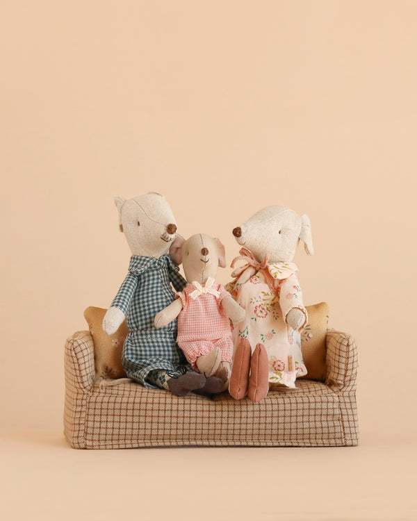 Three stuffed animals from the Maileg Grandparents & Me Set—a Grandma and Grandpa Mouse in charming patterned outfits alongside a Little Sister Mouse in pink overalls—sit together on a plaid sofa against a beige background, evoking the warmth of cozy family gatherings.