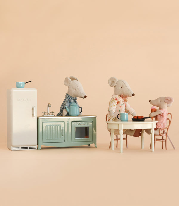 Three Maileg mice, including Grandma and Grandpa, gather at a table with food and cups beside the charming Maileg Kitchen Set, which features a miniature fridge, stove, and kettle, all set against a beige background. Their cozy world comes to life with delightful Maileg accessories and mouse-sized furniture from the Maileg Kitchen Set with Grandma & Grandpa.