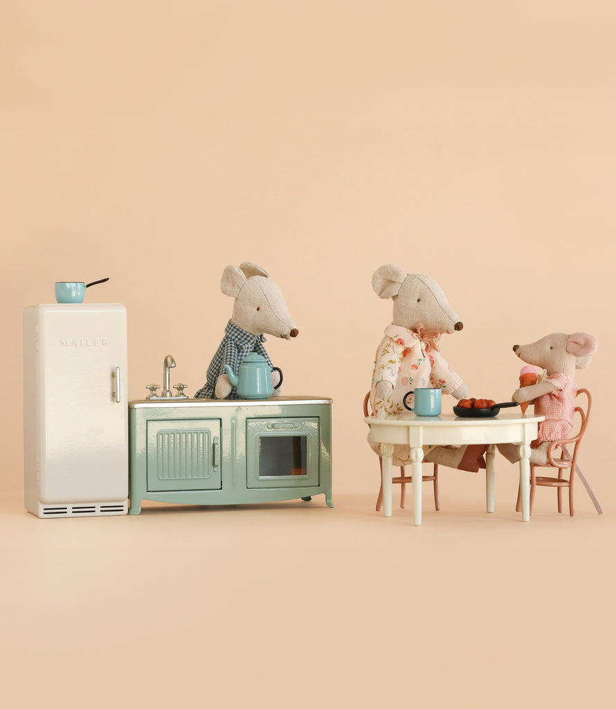 Three Maileg mice, including Grandma and Grandpa, gather at a table with food and cups beside the charming Maileg Kitchen Set, which features a miniature fridge, stove, and kettle, all set against a beige background. Their cozy world comes to life with delightful Maileg accessories and mouse-sized furniture from the Maileg Kitchen Set with Grandma & Grandpa.