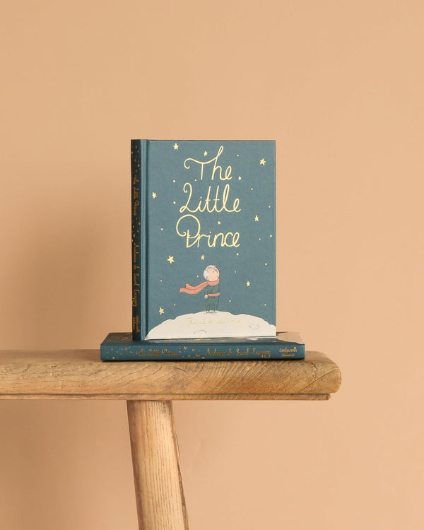 A copy of "The Little Prince | Collector's Edition | Hardcover" stands upright on a wooden bench, with another copy lying beneath it against a solid light beige background.