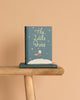 A copy of "The Little Prince | Collector's Edition | Hardcover" stands upright on a wooden bench, with another copy lying beneath it against a solid light beige background.