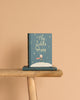 A copy of "The Little Prince | Collector's Edition | Hardcover" stands upright on a wooden bench, with another copy lying beneath it against a solid light beige background.