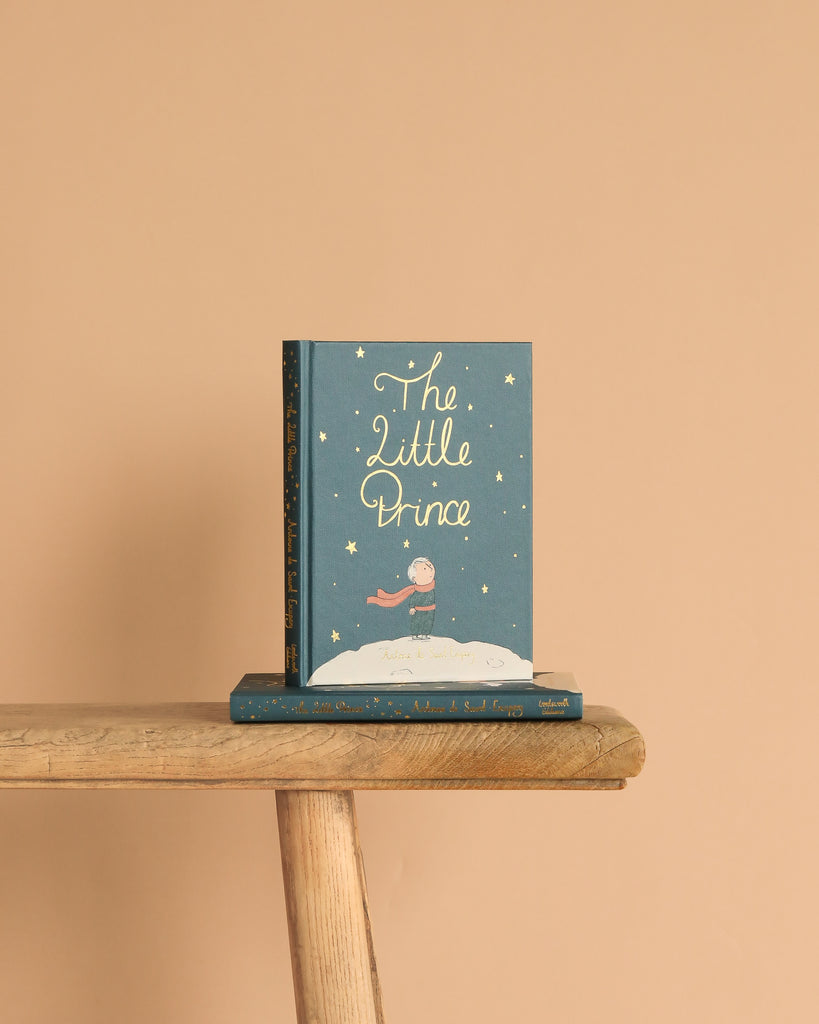 A copy of "The Little Prince | Collector's Edition | Hardcover" stands upright on a wooden bench, with another copy lying beneath it against a solid light beige background.