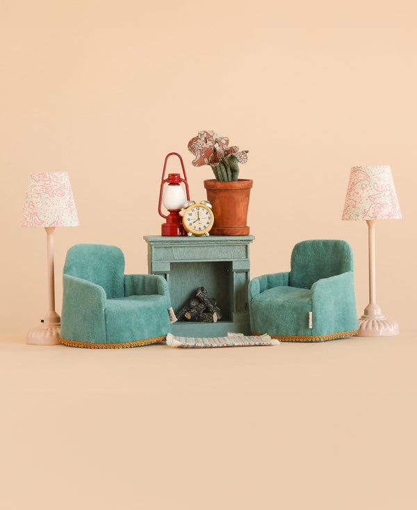 Transform your dollhouse with the Maileg Family Room Set, featuring two small teal armchairs from Maileg’s charming mouse furniture collection, arranged in front of a miniature fireplace decorated with delightful Maileg accessories such as a potted plant, lantern, and mantel clock. Two pink lamps frame the cozy scene against a soft beige background—an ideal addition to elevate your dollhouse furniture collection.