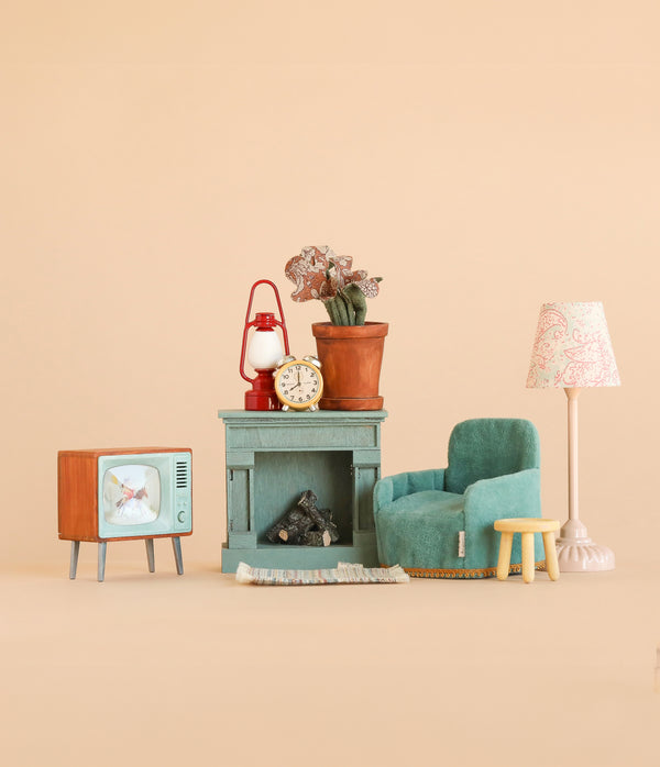 Bring a cozy touch to your miniature collection with the Maileg Family Room Set, featuring dollhouse accessories that include a teal armchair, small yellow stool, vintage TV, fireplace, rug, potted plant, clock, red lantern, and lamp. Perfect for creating charming scenes with Maileg or Maileg mouse furniture against a plain beige background.