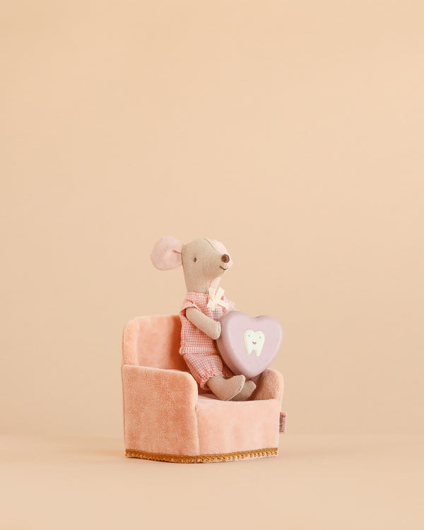 A Maileg Little Sister with Tooth Box Set mouse, dressed in pink, sits on a Maileg Soft Folding Chair and holds a purple heart-shaped tooth pillow against a plain beige background.
