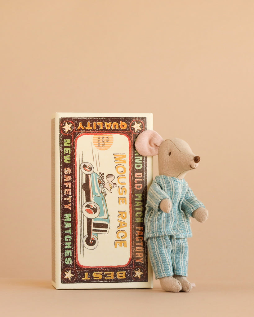 Maileg Big Brother, Mouse In Matchbox is a charming soft fabric toy featuring a small mouse dressed in blue pajamas, standing beside a vintage-style matchbox adorned with an illustration of a car race and the words “Mouse Race” on the cover. Set against a solid beige background, this delightful collectible is perfect for imaginative play and collectors alike.