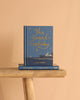 A blue hardcover Collector's Edition of "The Great Gatsby" stands upright on a wooden bench with another copy beneath it, set against a beige background. The cover displays gold stars and a seaside scene, highlighting this iconic novel.