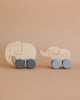 Two wooden toy elephants on wheels, designed to be free of harmful chemicals, are shown against a beige background. The larger elephant is in front with blue wheels, while the smaller one follows, holding the larger elephant’s tail with its trunk. These charming Wooden Mom and Baby Elephant Push Toy Set helps develop fine motor skills.