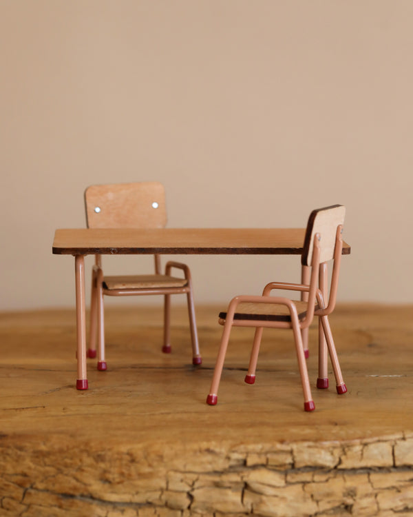 The Maileg School Table & Chair Set For Mice in Dark Powder features two small chairs and a table made of beech wood and metal, resting on a textured wooden surface. The chairs have light brown seats and backrests, with thin pinkish legs and red tips. The matching table showcases exquisite handmade color details.