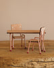 The Maileg School Table & Chair Set For Mice in Dark Powder features two small chairs and a table made of beech wood and metal, resting on a textured wooden surface. The chairs have light brown seats and backrests, with thin pinkish legs and red tips. The matching table showcases exquisite handmade color details.