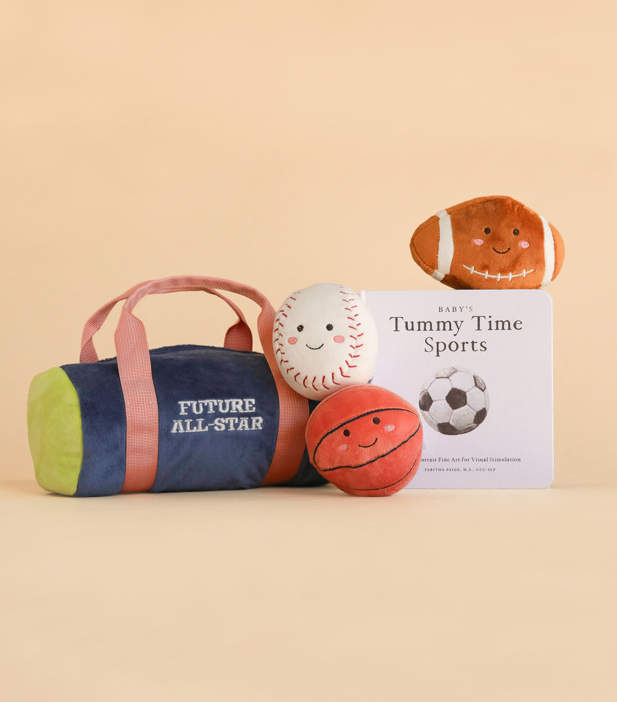 Plush sports toys from the “Future All-Star Baby Gift Set” and a book titled “Baby’s Tummy Time Sports” are arranged on a beige background. The set includes plushies shaped like a football, baseball, basketball, and rattle balls, along with a blue bag labeled “My First Sports Bag: Future All-Star.”.