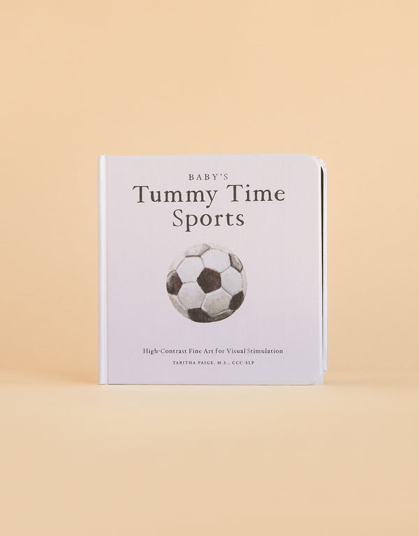 A high-contrast baby book, “Baby’s Tummy Time Sports,” features a bold black and white soccer ball illustration on the cover—a perfect choice for visual stimulation during tummy time. The book stands upright against a beige background.