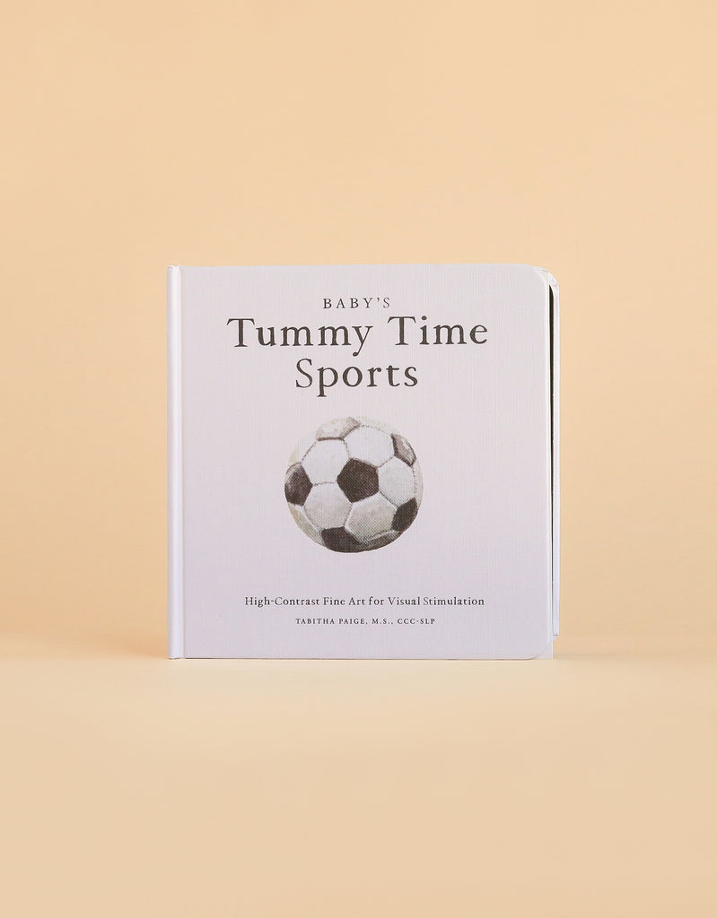 A high-contrast baby book, “Baby’s Tummy Time Sports,” features a bold black and white soccer ball illustration on the cover—a perfect choice for visual stimulation during tummy time. The book stands upright against a beige background.