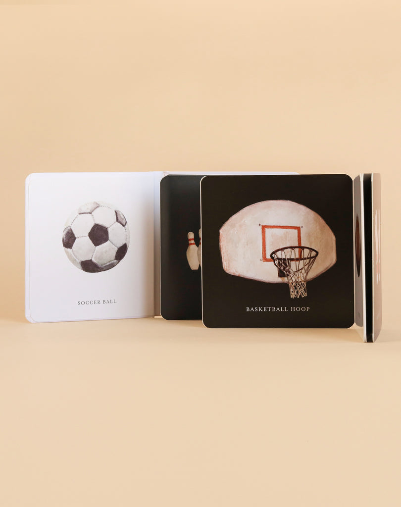The “Baby’s Tummy Time Sports” book is shown open, featuring high-contrast illustrations of a soccer ball, bowling pins, and a basketball hoop on different pages against a beige background—ideal for visual stimulation during tummy time.