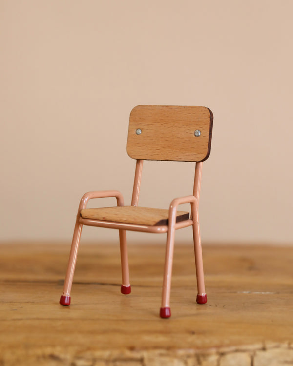 A small wooden Maileg Mouse Size School Chair in dark powder, featuring pink metal legs with red tips, stands on a wooden surface against a beige background. It has a straight backrest, armrests, and charming hand-painted details enhancing its whimsical appeal.