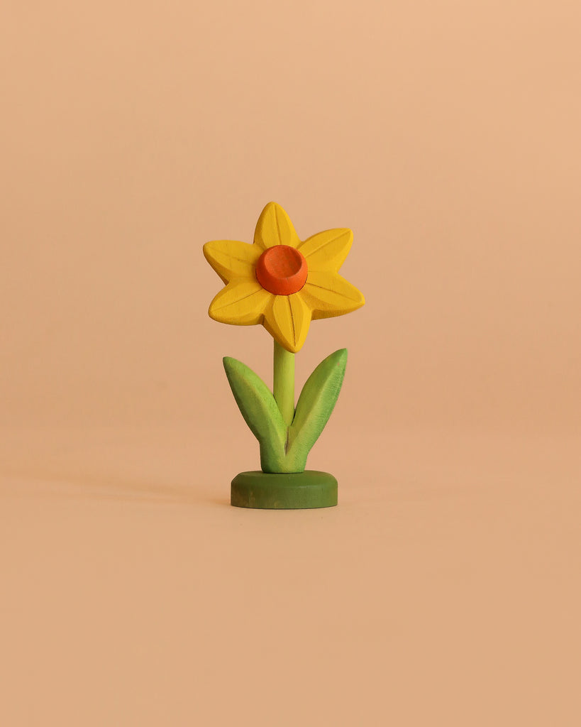 The Bumbu Handmade Wooden Flower Festive Daffodil features a yellow bloom with an orange center and green stem, standing upright on a light beige background—ideal for imaginative play or as a charming display in celebration rings.