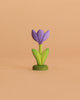 The Bumbu Handmade Wooden Flower Festive Crocus features a small purple flower with green leaves and stem, standing upright on a beige background—ideal as a Montessori toy or to add charm to a celebration ring.