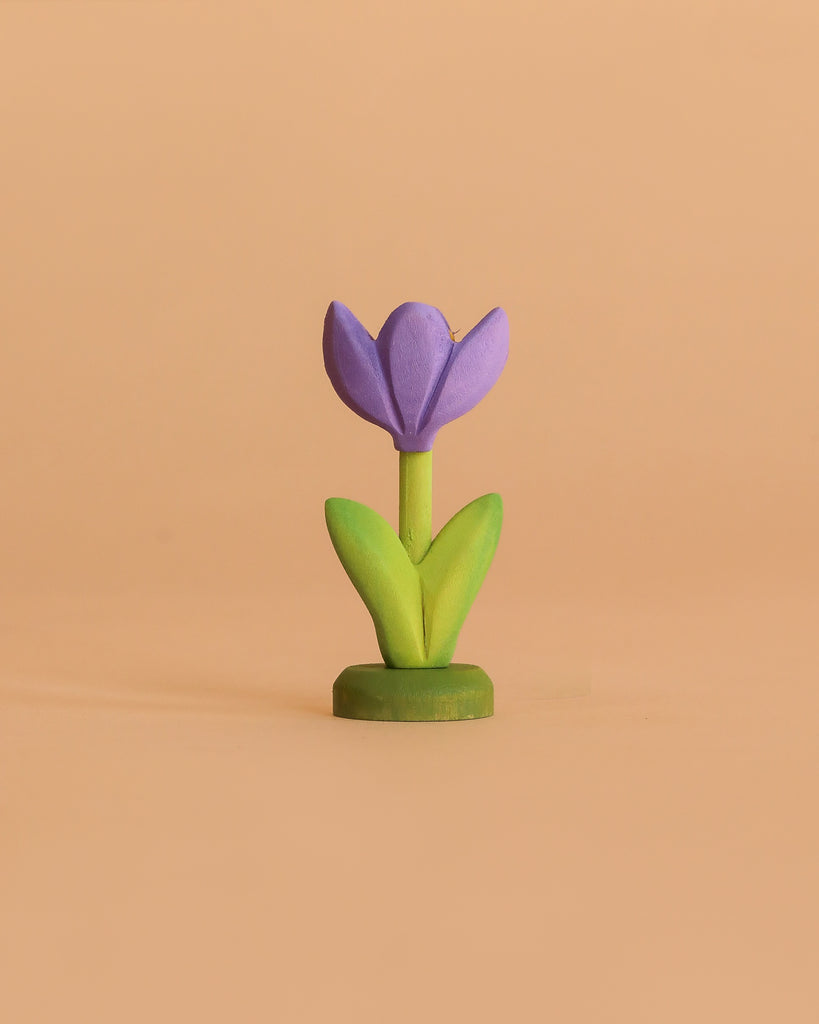 The Bumbu Handmade Wooden Flower Festive Crocus features a small purple flower with green leaves and stem, standing upright on a beige background—ideal as a Montessori toy or to add charm to a celebration ring.